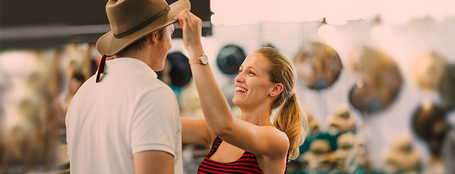 A woman is standing and smiling, putting a wide brim hat on a man standing opposite her 