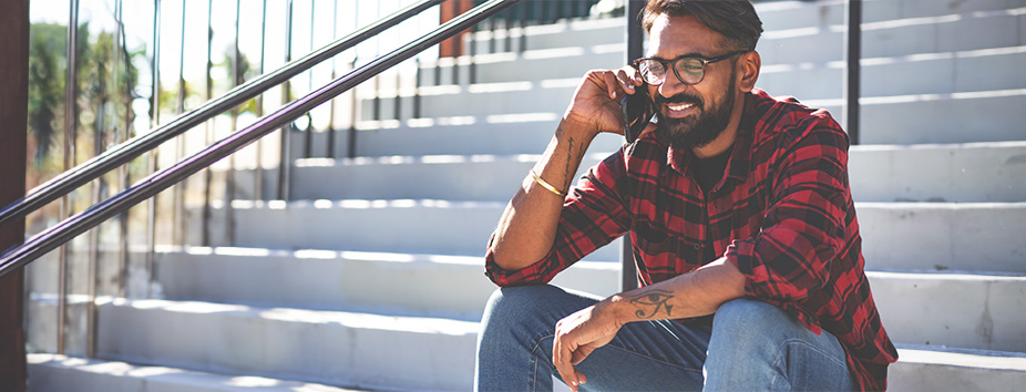 A young man with dark skin tone is sitting on a flight of steps outdoors. He's talking on his mobile phone and smiling. 